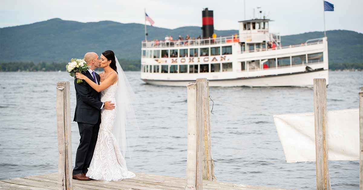 A couple kisses on a dock outside the Boathouse, with a steamboat in the background.